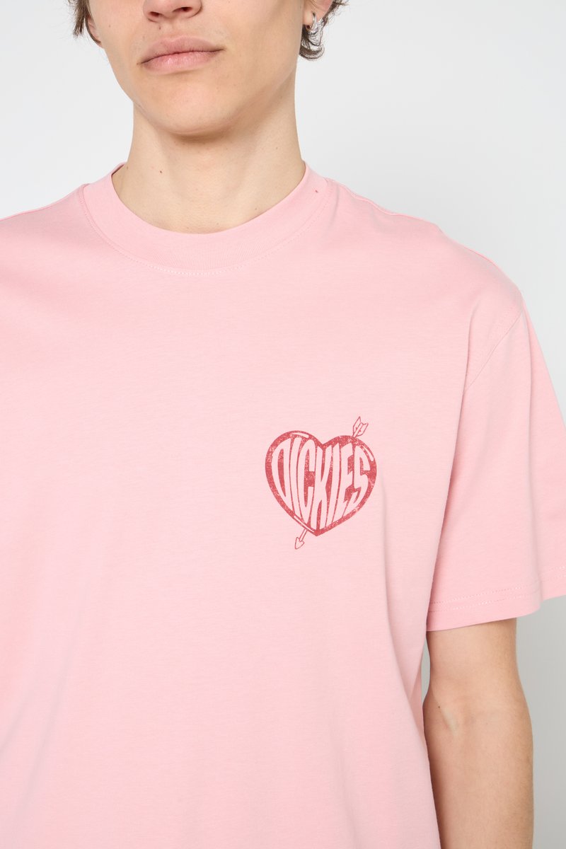 Homme portant un t-shirt rose clair avec un graphique en forme de cœur rouge et le texte "Dickies" traversé par une flèche sur la poitrine gauche.