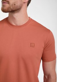 Trumpas rankovės terakotos marškinėliai, pagaminti iš lygaus audinio, su mažu kvadratiniu logotipo ženklu kairėje krūtinės pusėje ir apvalia iškirpte.