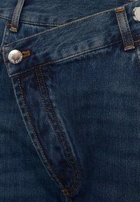 Dettagli della tasca frontale di un paio di jeans in denim blu con cuciture arancioni e chiusura a bottone argentato.