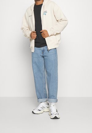 Felpa con zip crema sopra una t-shirt nera, abbinata a jeans azzurri con risvolto e sneakers bianche con accenti neri.