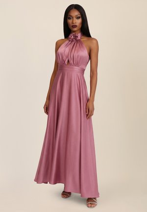 Frau steht in einem bodenlangen, staubrosafarbenen Neckholder-Kleid mit einer Rosetten-Ausschnitt und einem plissierten Oberteil, kombiniert mit silbernen, Riemchen-Absätzen.