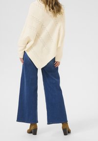 Beige stickad poncho med ett diagonalt mönster och öppna detaljer, bärs över blå vida jeans och bruna ankelstövlar med klumpig klack.