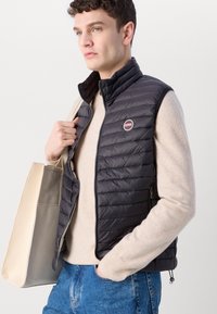 Gilet nero trapuntato con colletto alto, chiusura con zip e tasche laterali. Indossato sopra un maglione beige, abbinato a jeans blu e una borsa a tracolla beige.