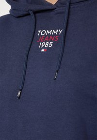 Felpa blu navy con cappuccio caratterizzata da cordini, con testo ricamato bianco e rosso: "TOMMY JEANS 1985" e un piccolo accento di logo. Texture morbida.