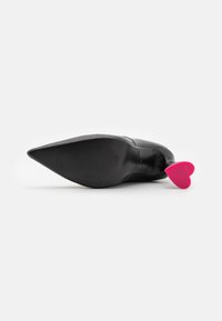 Zapato de tacón alto negro apuntado tumbado de lado con un tacón en forma de corazón rosa brillante, sobre un fondo blanco.