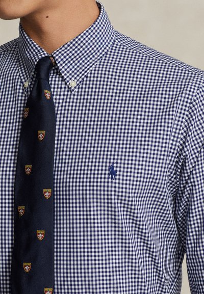 Blau-weiß kariertes Hemd mit Kragen und einer navyblauen Krawatte mit kleinen Wappenschilden, getragen von einer Person mit sichtbarem Hals und Kinn.