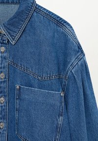 Chemise en denim bleu avec fermeture à boutons, col pointu et poche poitrine avec coutures visibles et rivets métalliques sur fond clair.