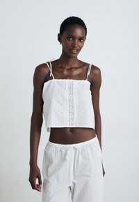 Top alb cu croptop, cu guler elastic și bretele ajustabile, decorat cu broderie florala și detalii de dantelă intricate. Asortat cu pantaloni albi brodați.