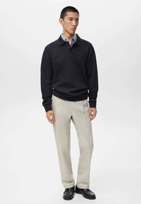 Suéter de punto negro con cuello, combinado con pantalones beige claros. Modelo que lleva zapatos negros, de pie sobre un fondo blanco liso.