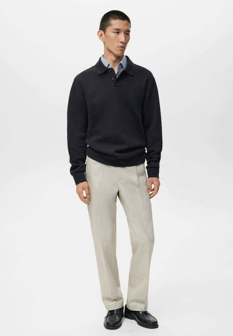Pull noir en tricot avec un col, associé à un pantalon beige clair. Modèle portant des chaussures noires, se tenant devant un fond blanc uni.
