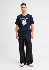 Navy blauwe T-shirt met een cartoon kerstman in een roze opblaasband en de tekst "Kerstman is druk", gecombineerd met losse donkere spijkerbroek en witte sneakers.