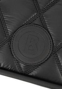 Schwarzer gestepptes Stoff mit glänzender Textur, versehen mit einem runden Lederpatch, auf dem der Buchstabe "B" eingeprägt ist. Sichtbare Steppnähte.
