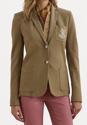Kvinde iført en brun blazer med to forlommer og broderet logo på brystet, kombineret med lyserøde bukser og et flerfarvet tørklæde.