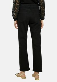 Jeans droits en denim noir avec deux poches arrière, des détails subtils sur les côtés, et une taille haute. Associés à des sandales dorées.