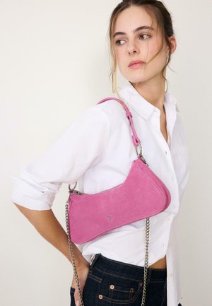 Woman wearing white shirt and dark jeans carries small pink suede shoulder bag with silver chain strap.