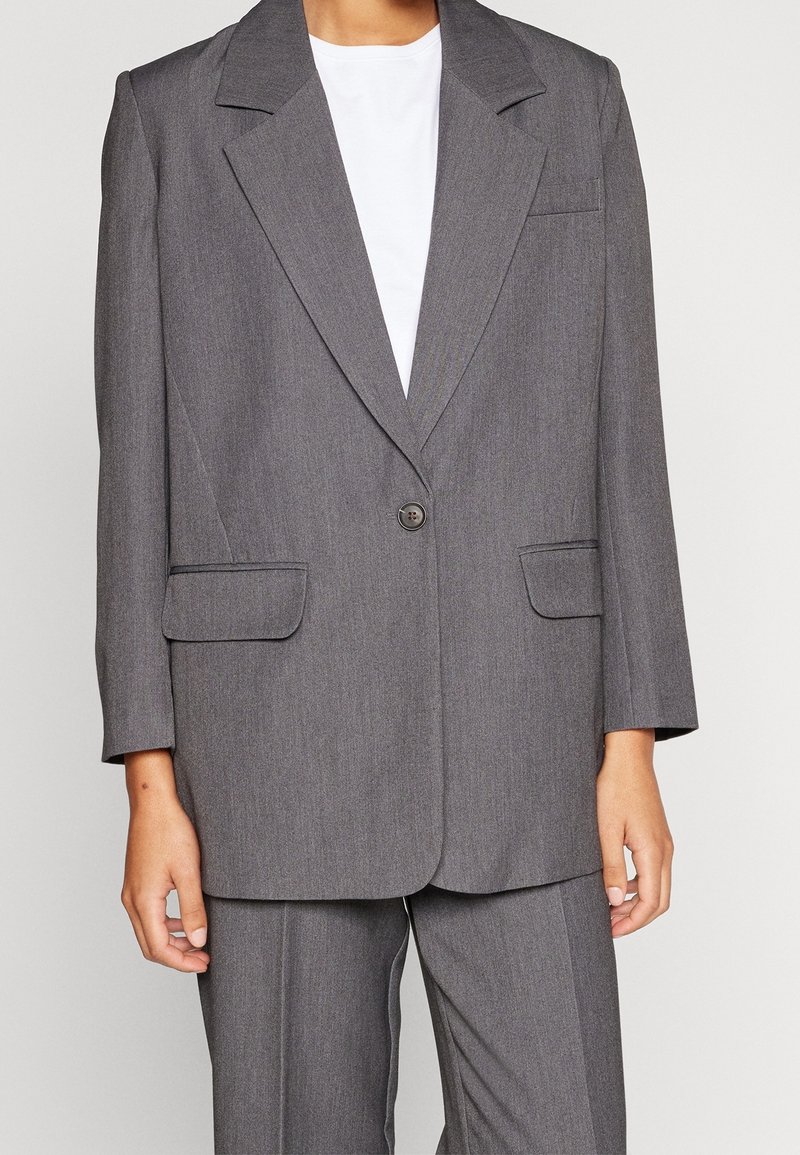 Blazer grigio su misura realizzato in un tessuto texturizzato, con chiusura a un bottone, rever affilati e due tasche frontali.