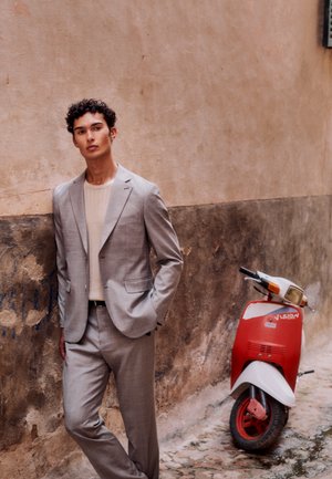 Tânăr îmbrăcat într-un costum gri deschis și o cămașă bej, stând cu mâinile în buzunare lângă o scuter vintage roșu și alb, în fața unui perete texturat.