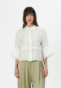 Blouse blanche avec des détails frangés au col et aux manches ; coupe ample, tissu léger ; associée à un pantalon vert olive.