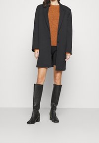 Svart lång kappa över en orange stickad tröja, i kombination med svarta shorts och knähöga läderstövlar. Snygg design och strukturerad passform.