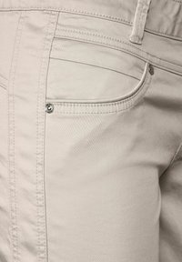 Beiga bomullbyxor med en slät textur, med sömmar på bakfickorna och metallknappdetaljer. Närbild från sidan.