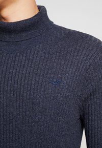 Granatowy sweter z golfem w prążki wykonany z miękkiej dzianiny. Posiada małe, ciemne logo na piersi oraz dopasowaną sylwetkę.