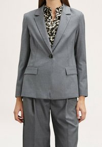 Blazer gris sur mesure avec un bouton unique, deux poches avant et des épaules structurées, associé à une chemise à imprimé léopard en dessous.