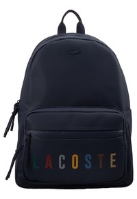 Laivaston sininen reppu, jossa on etutaskun vetoketju, monivärinen "LACOSTE"-teksti ja pieni krokotiililogo taskun yläpuolella.