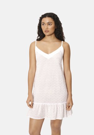 Mujer con cabello largo y rizado, vestida con un vestido blanco de ojales sin mangas, con tirantes finos y un dobladillo con volantes, de pie frente a un fondo liso.