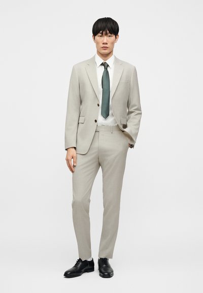 Ανοιχτό γκρι κοστούμι με μονόπετο σακάκι, παντελόνι στενής γραμμής, λευκό πουκάμισο και πράσινη γραβάτα με σχέδια. Μαύρα επίσημα παπούτσια ολοκληρώνουν την εμφάνιση.