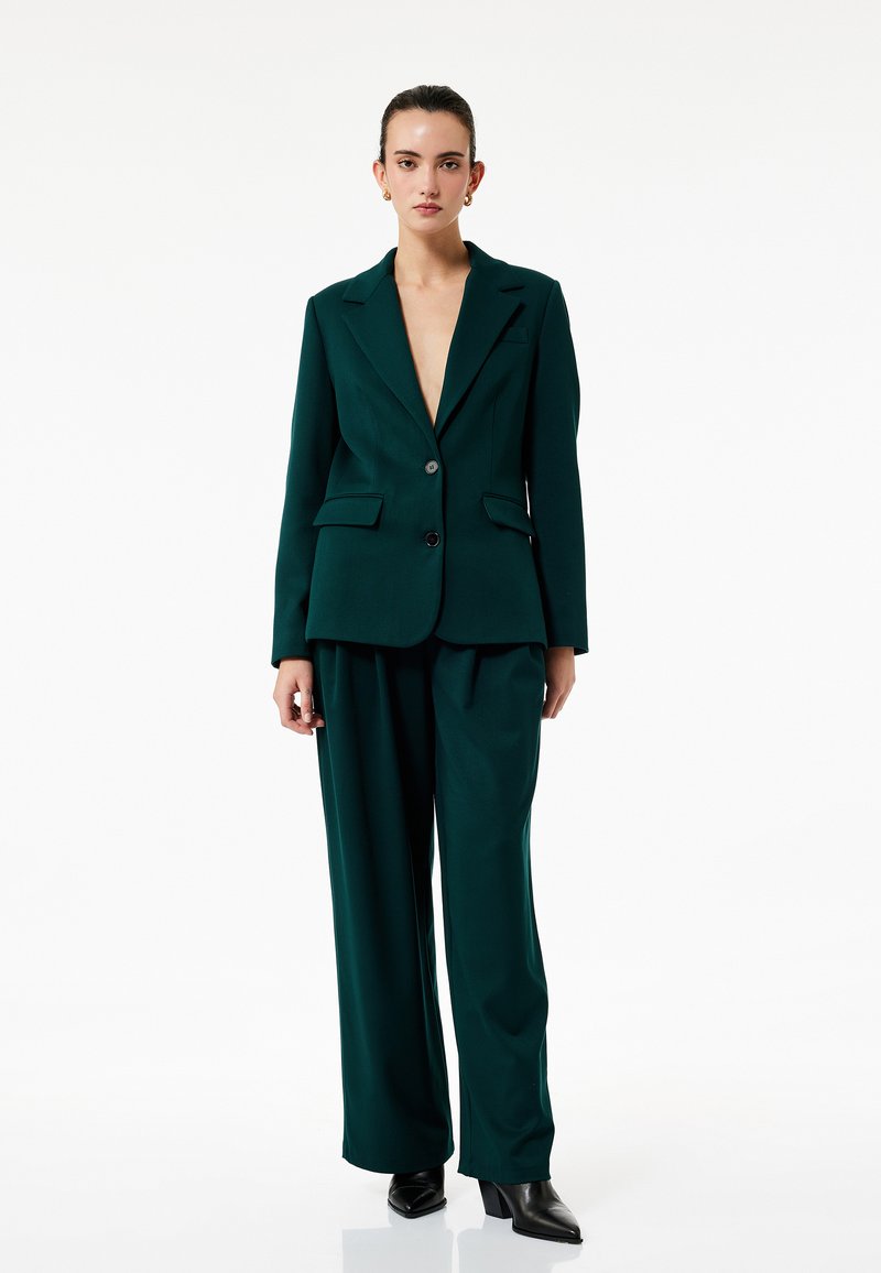 Skovgrøn blazer med spidse revers og to knapper, parret med løstsiddende, matchende bukser. Glat stof og minimalistisk design.