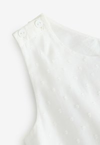 Prenda blanca sin mangas con pequeños puntos elevados y texturizados y dos botones blancos en la tira del hombro.