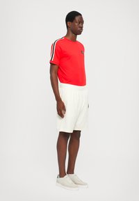 Rød kortermet T-skjorte med svarte og hvite sidestriper og logo, kombinert med hvite shorts med elastisk liv og lysegrå sneakers.