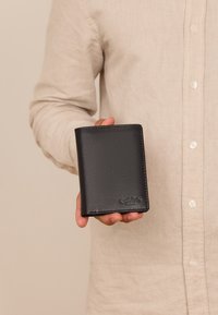 Černá kožená peněženka s hladkým povrchem, skládací design a vyražené logo. Držena v ruce na světlejším pozadí.