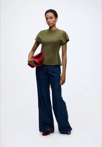 Γυναίκα με ελιά πράσινο κοντομάνικο τοπ, φαρδιά σκούρα μπλε παντελόνια με κουμπιά στο πλάι, κρατώντας μια μεγάλη κόκκινη τσάντα φάκελο, στέκεται μπροστά σε λευκό φόντο.