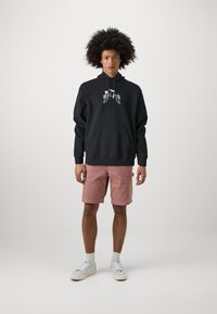 Sudadera negra con bolsillo frontal y diseño gráfico blanco, combinada con pantalones cortos rosas y zapatillas blancas con accents grises.