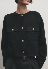 Svart kort tröja med två framfickor, silverfärgade knappstängningar och ribbat mönster. Har rund halsringning och långa ärmar.