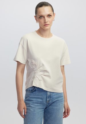 Camiseta de algodón de color crema, de manga corta, con bordado floral en el lado izquierdo. Textura suave, corte relajado y diseño de cuello redondo.