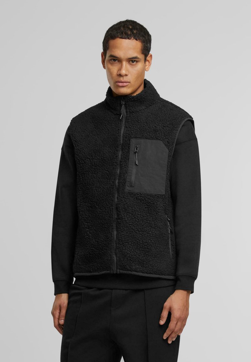 Schwarze Fleece-Weste mit hohem Kragen, durchgehendem Reißverschluss und einer kontrastierenden schwarzen Stofftasche. Verfügt über einen strukturierten Körper und seitliche Reißverschlüsse.