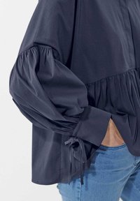 Navyblaues Hemd mit Knopfleiste und voluminösen Puffärmeln, die an den Manschetten mit Bändern gerafft sind, sowie einem ausgestellten Saum. Glatte Baumwollstoffstruktur.