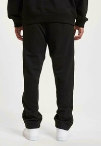 Svart joggebukse laget av mykt stoff med en avslappet passform, med sidelommer, elastisk midje og glidelåser ved anklene. Hvite sneakers.
