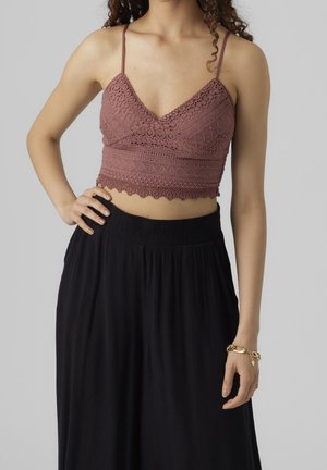 Vrouw draagt een mauve gehaakt cropped topje met spaghettibandjes en een loszittende zwarte broek met hoge taille, hand op de heup, gouden armband om de pols.