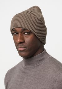 Brun ribbad stickad mössa med en spetsig topp, bärs över en ljusgrå högkragad tröja. Mössans textur är slät och något elastisk.