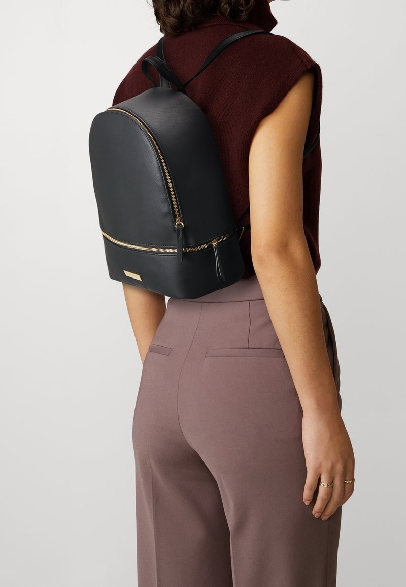 Personne portant un haut sans manches rouge foncé et un pantalon mauve avec un sac à dos en cuir noir avec des fermetures éclair dorées et une petite étiquette à l'avant.