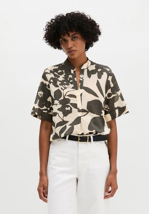 Kobieta o kręconych włosach, ubrana w beżową i czarną kwiecistą bluzkę z krótkim rękawem, włożoną do białych spodni z czarnym paskiem.