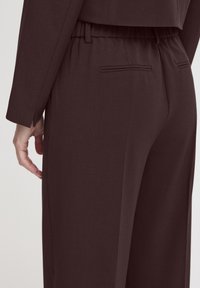Brown tailored trousers with a smooth texture, elastic waistband, and two rear pockets, featuring subtle stitching details and a straight fit.