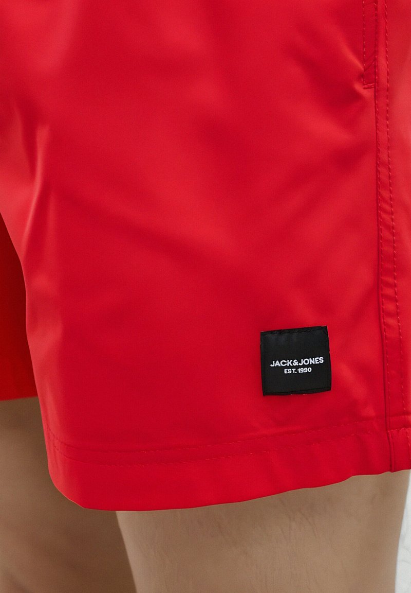 Piros úszónadrág sima, könnyű anyagból. Fekete címke található a szegélyén, amin ez áll: "JACK & JONES EST. 1990".
