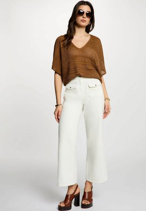 Donna che indossa una maglia marrone testurizzata a maniche corte, pantaloni bianchi a vita alta e gamba larga, sandali marroni con plateau e occhiali da sole scuri, in piedi su sfondo bianco.