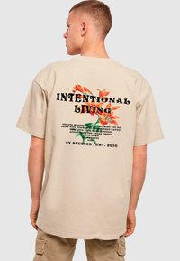 Beige oversized t-skjorte med blomstertrykk og typografi på baksiden, med teksten "INTENTIONAL LIVING" i fete svarte bokstaver.