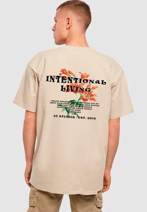 Tricou oversized bej cu imprimeu floral și tipografie pe spate, cu expresia "VIEȚUIRE INTENȚIONATĂ" în litere negre îndrăznețe.