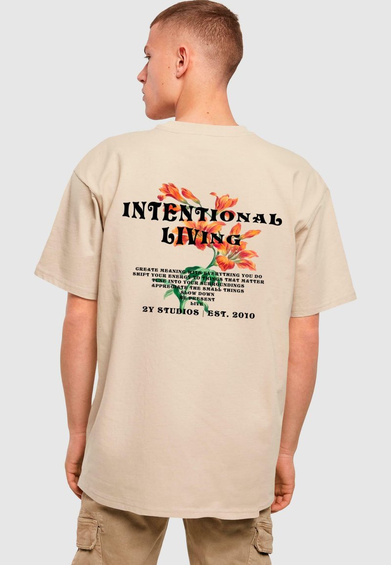 Beige oversized t-skjorte med blomstertrykk og typografi på baksiden, med teksten "INTENTIONAL LIVING" i fete svarte bokstaver.